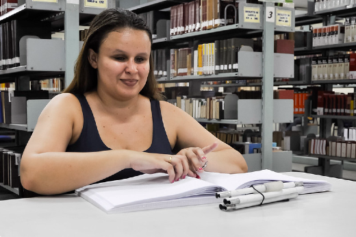 Imagem: Mulher branca de cabelos pretos sentada em mesa dentro de biblioteca fazendo leitura em braille de livro