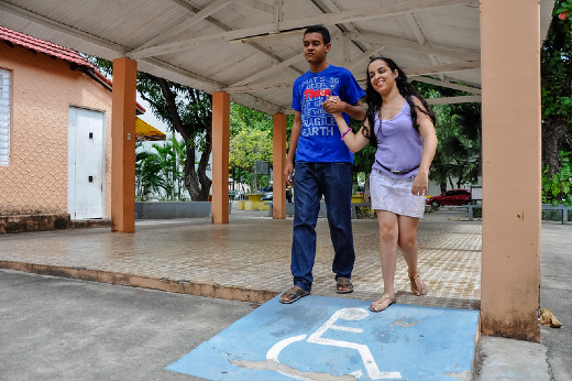 Imagem: estudante apoia pessoa com deficiência visual a caminhar no campus