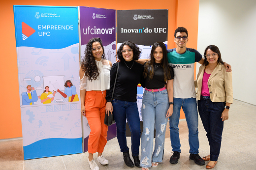 Imagem: grupo de estudantes, homens e mulheres, em pé na frente de banner do evento