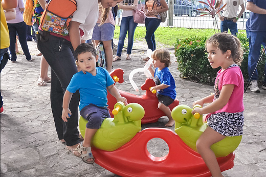 Imagem: crianças brincam em parquinho em ambiente aberto com adultos ao redor.