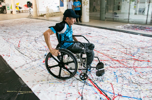 Imagem: Homem em cadeira de rodas sobre papel branco, pintado com várias cores (Foto: Ribamar Neto/UFC Informa)
