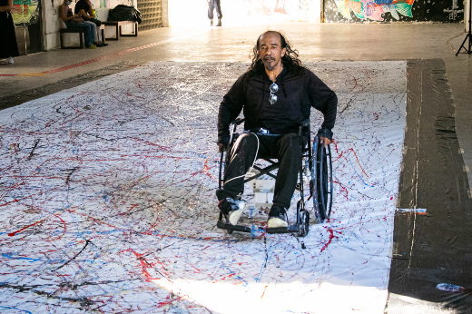 Imagem: Em primeiro plano, homem em cadeira de rodas sobre um grande papel branco; da cadeira de rodas sai tinta colorida que pinta o painel (Foto: Ribamar Neto/UFC Informa)