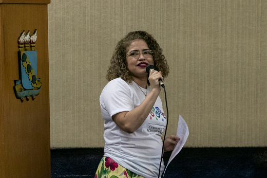 Imagem: A bibliotecária Cléo Sousa fala ao microfone durante evento da Secretaria de Acessibilidade no auditório da Reitoria. Ela tem cabelos cacheados e traja camiseta branca. (Foto: Ribamar Neto/UFC Informa)