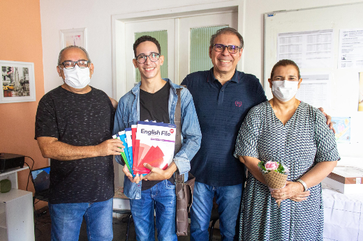 Imagem: Foto mostra quatro pessoas de frente. Da esquerda para direita: um professor de camisa preta; um jovem estudante mostrando a coleção de livros didáticos que recebeu como premiação pelo concurso; o coordenador da Casa de Cultura Britânica, homem branco de camisa azul; e a vice-coordenadora, que usa vestido e máscara (Foto: Gutierrez Reges/UFC Informa)