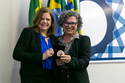 A secretaria das Relações Internacionais do Ceará, Roseane Oliveira de Medeiros, de blazer preto e camisa azul, posa com sua medalha ao lado da vice-reitora da UFC, Diana Azevedo, de blazer preto, camisa estampada  e cabelo curto cacheado