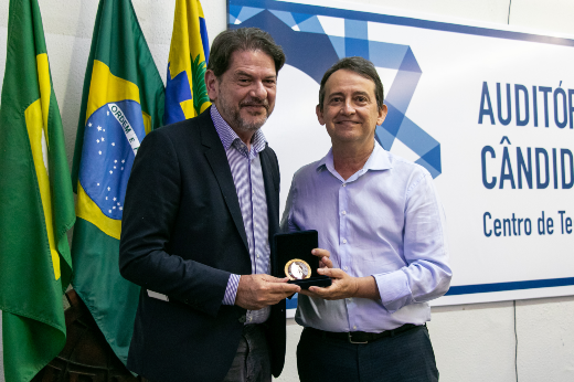 Senador Cid Gomes, vestindo bazer preto, calça jeans e camisa listrada clara, recebe medalha das mãos do professor Almir Holanda, que veste calça social escura e camisa clara