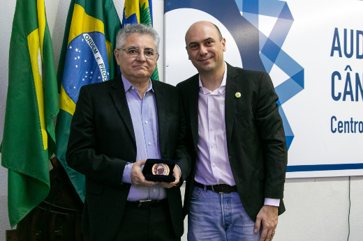 Professor Bruno Vieira, calvo, de calça jeans e bazer, posa ao lado do professor Francisco de Assis de Souza Filho, de óculos, paletó e que segura uma medalha em uma caixa