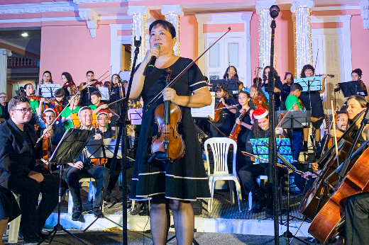 professora do ICA aparece segurando um violino, de vestido preto e cabelo preso, à frente da Orquestra e da Camerata de Cordas da UFC, com músicos sentados segurando seus instrumentos, vestidos de preto