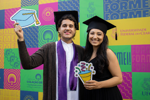 Imagem: formando e amiga posam para foto em frente a backdrop com marca da colação de grau em Fortaleza