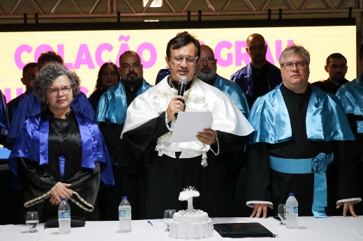 Imagem: da esquerda para a direita, aparecem a vice-reitora Diana Azevedo, o reitor Custódio Almeida, segurando folhas de papel enquanto lê o discurso, e o pró-reitor de graduação Davi Romero. Todos usam suas respectivas vestes talares