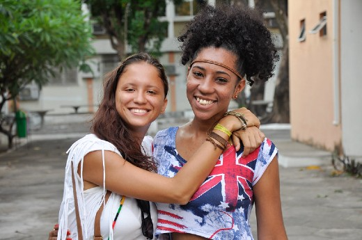 Imagem: duas mulheres uma negra com blusa colorida e cabelo crespo preso e outra branca com cabelos longos castanho abraçadas 