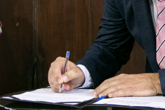 Imagem: Close-up de uma mão segurando uma caneta esferográfica com ponta azul, no momento de assinar um documento sobre uma mesa. Ao lado, vê-se parte da outra mão apoiada no papel. A pessoa veste camisa branca e paletó azul-escuro