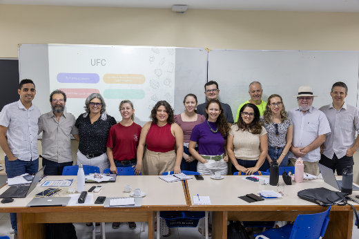 Imagem: Esta imagem mostra um grupo de 14 pessoas, compostas por homens e mulheres adultos, posando para uma foto em uma sala de aula ou reunião. Eles estão de pé, lado a lado, atrás de duas mesas grandes organizadas em forma de U, sobre as quais há cadernos, garrafas, celulares, canetas, papéis e notebooks — sugerindo um ambiente de trabalho ou reunião acadêmica.  Ao fundo, há um projetor exibindo um slide com o título "UFC", o que indica que o evento pode estar relacionado à Universidade Federal do Ceará. O fundo é composto por quadros brancos e paredes claras. A maioria das pessoas está sorrindo, aparentando um clima amigável e colaborativo.
