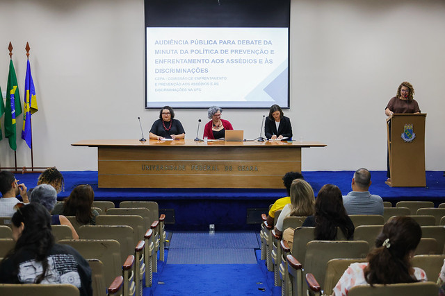 Imagem: três mulheres sentadas em uma mesa e outra mulher falando no púlpito do auditório da Reitoria