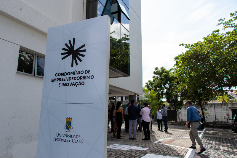 Imagem: A imagem mostra a entrada de um prédio moderno com uma placa grande em destaque. Na placa, lê-se: "CONDOMÍNIO DE EMPREENDEDORISMO E INOVAÇÃO" "UNIVERSIDADE FEDERAL DO CEARÁ" Abaixo do texto está o brasão da universidade. Na frente do prédio, há várias pessoas reunidas, possivelmente participando de um evento. O ambiente é urbano, com árvores ao redor e piso de blocos intertravados. A fachada do edifício é branca, com janelas espelhadas, refletindo a vegetação ao redor. A cena ocorre durante o dia, sob luz natural.
