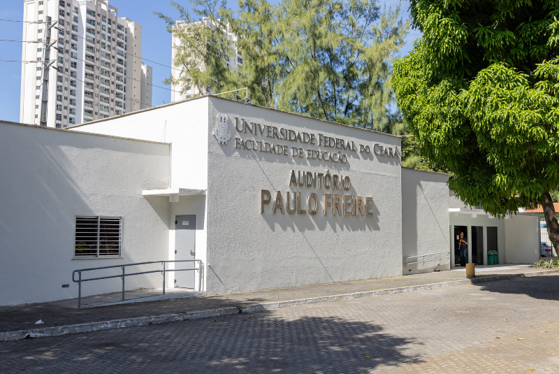 Imagem: A imagem mostra a fachada do Auditório Paulo Freire, localizado na Faculdade de Educação da Universidade Federal do Ceará (UFC). A construção é de cor clara, com letras metálicas em relevo destacando o nome da instituição e do auditório. À esquerda, há uma rampa de acessibilidade ao lado de uma pequena janela. À direita, há uma entrada maior com portas de vidro, parcialmente abertas, por onde se vê uma pessoa no interior. Ao fundo, vemos edifícios residenciais altos, árvores e céu claro, sugerindo que o local está situado em uma área urbana bem arborizada. Há também uma grande árvore com sombra à direita, que contrasta com o cimento do pátio frontal. O ambiente é limpo e bem conservado.