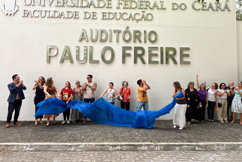 Imagem: A imagem mostra um grupo de pessoas participando de uma cerimônia de inauguração ou renomeação em frente a um prédio da Universidade Federal do Ceará (UFC), especificamente da Faculdade de Educação. A parede ao fundo exibe em letras grandes:  AUDITÓRIO PAULO FREIRE  As pessoas estão posicionadas em linha, algumas aplaudindo e sorrindo. Há um pano azul sendo retirado, indicando que o nome “PAULO FREIRE” acabou de ser revelado. A cena é de celebração e homenagem, provavelmente relacionada ao legado de Paulo Freire, importante educador e filósofo brasileiro. A presença de diversas pessoas, com expressões de alegria e admiração, sugere um evento institucional e simbólico de grande significado.