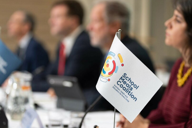 Imagem: A imagem mostra uma reunião formal com foco em uma pequena bandeira de mesa com o logotipo da School Meals Coalition. Abaixo do nome, lê-se o slogan: "Nutrition, Health and Education for Every Child" (Nutrição, Saúde e Educação para Toda Criança).  No fundo, desfocados, estão quatro participantes sentados à mesa, com roupas formais, indicando que se trata de um evento institucional ou conferência internacional. À direita da imagem, uma mulher com blusa bordô e colar amarelo está parcialmente em foco, prestando atenção no evento.  A mesa está organizada com papéis, tablets e copos d’água, reforçando o caráter sério e profissional do encontro. A presença da bandeira e do logotipo sugere que o encontro está relacionado a discussões sobre políticas públicas ou iniciativas globais voltadas à alimentação escolar infantil.