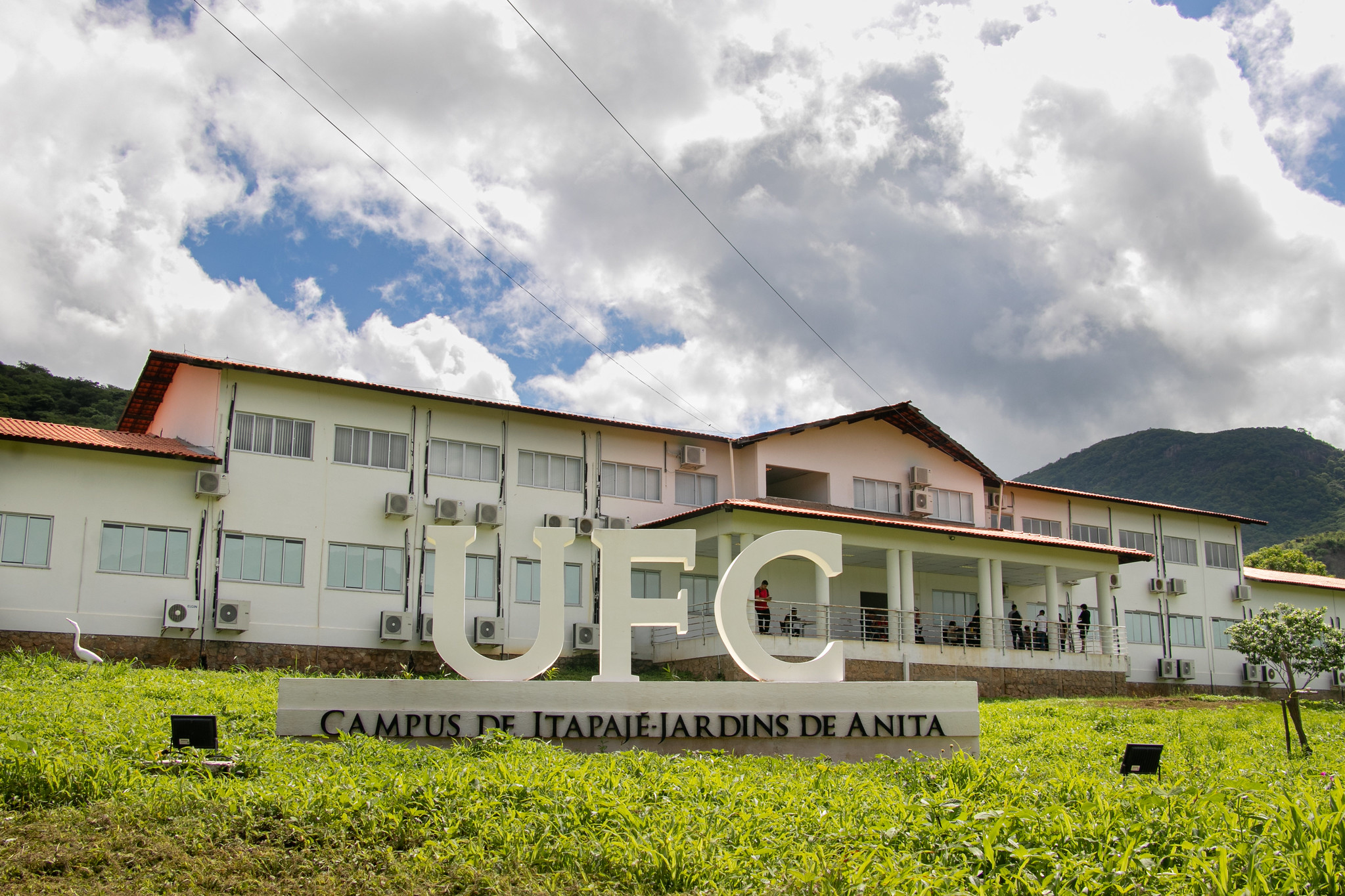 Imagem: Foto colorida de um prédio branco de dois andares com telhado de telhas vermelhas, pertencente à Universidade Federal do Ceará (UFC), Campus de Itapajé – Jardins de Anita. Em frente ao prédio, há um letreiro grande com as letras “UFC” em destaque e abaixo a identificação do campus. Algumas pessoas estão reunidas sob uma marquise no térreo. O céu está parcialmente nublado, e ao fundo vê-se uma paisagem com montanhas cobertas por vegetação verde.