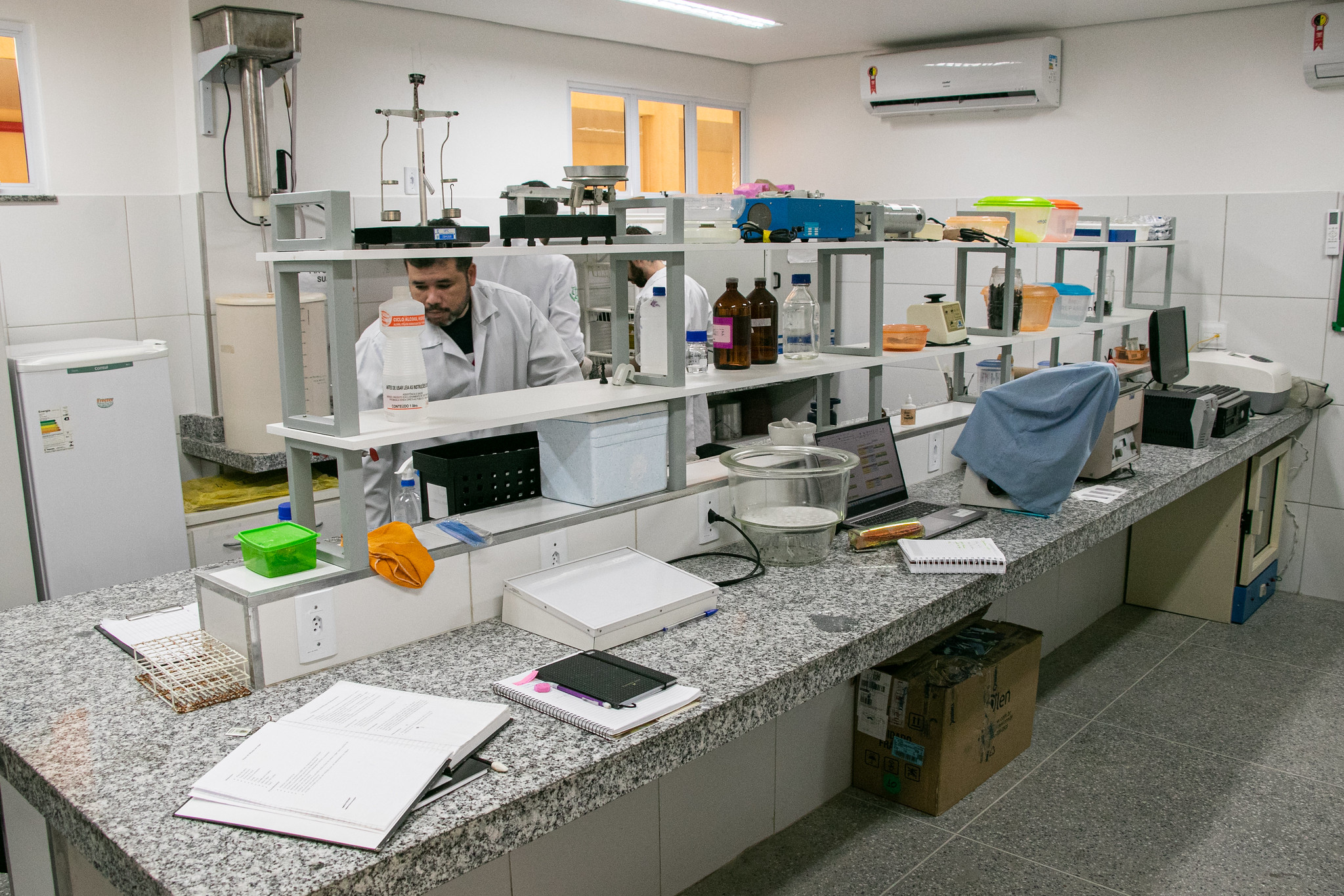 Imagem: Foto colorida de um laboratório moderno com bancadas de granito, localizado em um campus universitário. No centro da imagem, um homem com jaleco branco realiza atividades de pesquisa ao lado de outros colegas parcialmente visíveis. As bancadas estão cobertas por livros, cadernos, frascos com reagentes, equipamentos científicos e um notebook aberto. O ambiente é bem iluminado, possui paredes revestidas com azulejos brancos, janelas de vidro e dois aparelhos de ar-condicionado fixados na parte superior. O laboratório aparenta ser voltado para análises químicas ou biológicas.