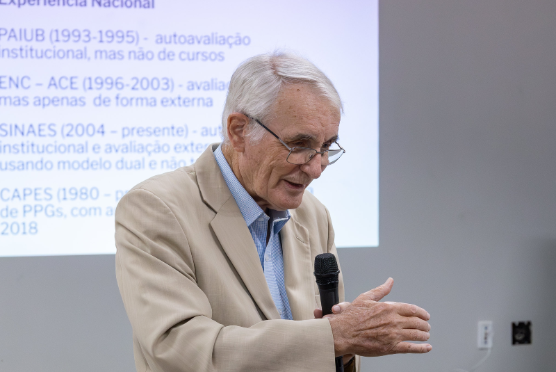 Imagem: A imagem mostra um senhor idoso realizando uma apresentação ou conferência. Ele está vestindo um terno bege claro com camisa azul e segura um microfone na mão direita. Seu corpo está levemente inclinado para frente, com a mão esquerda gesticulando, indicando que ele está explicando algo com ênfase e clareza. Ele também usa óculos de leitura, que estão posicionados na ponta do nariz, enquanto olha para baixo, provavelmente em direção ao público ou às próprias anotações.  Ao fundo, há uma projeção com texto em português, mencionando siglas como PAIUB, ENC-ACE, SINAES e CAPES, relacionadas a políticas de avaliação da educação superior no Brasil. Isso sugere que a palestra trata de temas ligados à avaliação institucional ou à educação superior.  O ambiente parece ser uma sala de aula ou auditório moderno, com paredes claras e equipamentos de projeção. A postura e expressão do palestrante demonstram experiência e domínio do tema abordado.