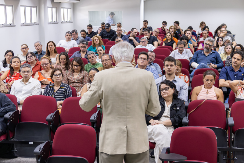 Imagem: A imagem mostra um seminário sendo realizado em uma sala de aula ou auditório. No primeiro plano, está um homem de cabelos grisalhos, vestindo um terno bege, visto de costas — ele parece ser o palestrante ou mediador da atividade. Ele está de pé, falando com o público, que está distribuído em cadeiras vermelhas com apoio para escrita.  A plateia está atenta, composta por homens e mulheres de diferentes idades, a maioria com expressões sérias ou concentradas, sugerindo que o tema abordado é de natureza acadêmica ou profissional. As pessoas estão bem-arrumadas, com roupas casuais ou sociais leves.  A sala é bem iluminada, com janelas ao longo da parede esquerda e uma cabine de vidro ao fundo, onde duas pessoas podem ser vistas, talvez operando equipamentos de som ou vídeo. O ambiente é formal e parece fazer parte de uma instituição de ensino ou centro de conferências.