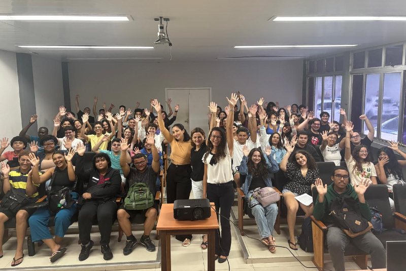 Imagem: A imagem mostra um grupo grande de estudantes reunidos em uma sala de aula ou auditório na unidade didático do Labomar (Instituto de Ciências do Mar da Universidade Federal do Ceará). Os alunos estão sentados em fileiras de cadeiras com braços de apoio, e todos estão voltados para a câmera, sorrindo e com as mãos levantadas, em um gesto de saudação ou entusiasmo coletivo.  Na frente da sala, há uma mesa com um projetor, e ao redor dela estão algumas pessoas em pé, que parecem ser professoras, monitoras ou organizadoras da atividade — todas sorrindo, reforçando o clima positivo do encontro.  A sala é bem iluminada, com grandes janelas laterais que deixam entrar luz natural, além de lâmpadas fluorescentes no teto. O ambiente sugere um momento de confraternização acadêmica, como o encerramento de um curso, uma recepção para calouros, uma palestra ou outro tipo de evento educativo. A diversidade dos participantes também indica um ambiente inclusivo e acolhedor.
