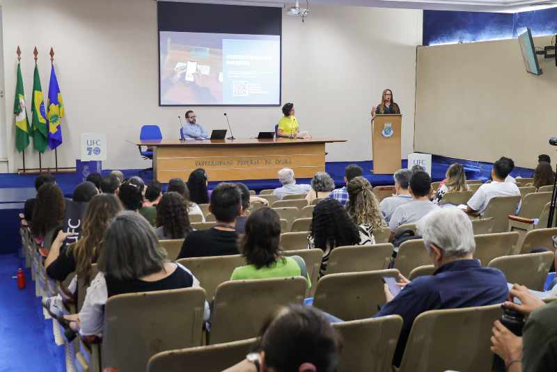 Imagem: A imagem mostra o 1º Encontro da Rede de Comunicadores da UFC (Universidade Federal do Ceará), realizado no auditório da Reitoria da instituição. O ambiente é formal e bem organizado, com diversas pessoas sentadas em poltronas bege, voltadas para o palco onde ocorre a apresentação. A plateia é composta por um público diverso, atento e engajado.  No palco, há três pessoas: uma mulher está em pé no púlpito, discursando; outra mulher, vestindo camiseta amarela, está sentada ao centro da mesa principal; e um homem de camisa azul está sentado ao lado esquerdo. Um telão ao fundo exibe uma apresentação com a legenda “Contatos da UFC Informa”, acompanhada de um QR Code. Ao lado do palco, há três bandeiras: do Brasil, do Ceará e da UFC.  O espaço é decorado com tons de azul e bege, com destaque para o nome “Universidade Federal do Ceará” gravado na mesa central. A presença de equipamentos como câmera e microfones indica que o evento pode estar sendo registrado ou transmitido.  O encontro é uma iniciativa institucional voltada à integração e capacitação dos profissionais de comunicação da universidade.