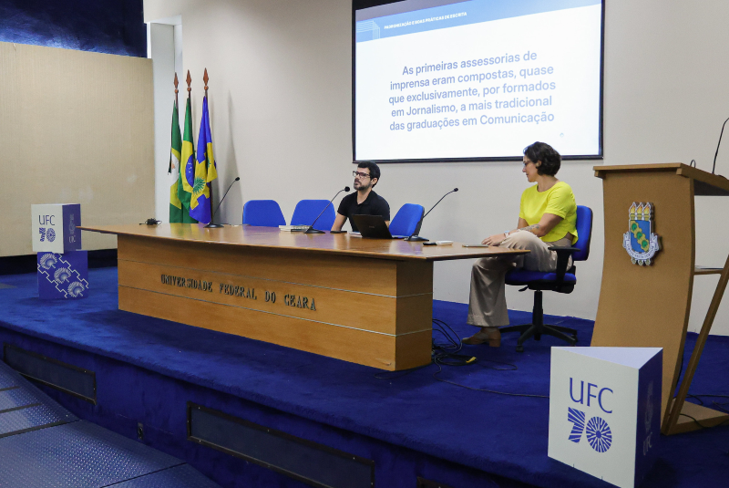 Imagem: A imagem retrata mais um momento do 1º Encontro da Rede de Comunicadores da UFC (Universidade Federal do Ceará), realizado no auditório da Reitoria. Dois palestrantes estão sentados à mesa principal, engajados em uma apresentação sobre a história e a formação das assessorias de imprensa.  No telão ao fundo, a projeção exibe o trecho: "As primeiras assessorias de imprensa eram compostas, quase que exclusivamente, por formados em Jornalismo, a mais tradicional das graduações em Comunicação", indicando uma reflexão sobre a evolução do perfil profissional na área da comunicação institucional.  Ambos os apresentadores utilizam microfones, e um deles está com um notebook aberto à sua frente. O ambiente é organizado, com o púlpito institucional à direita, decorado com o brasão da UFC, e as bandeiras oficiais (Brasil, Ceará e UFC) posicionadas à esquerda. O palco e o carpete são de cor azul, remetendo às cores institucionais da universidade. Também se destaca o logotipo dos 70 anos da UFC, visível em dois pontos do palco.  A imagem sugere um momento mais analítico do evento, com foco em aspectos históricos e profissionais da comunicação pública e universitária.