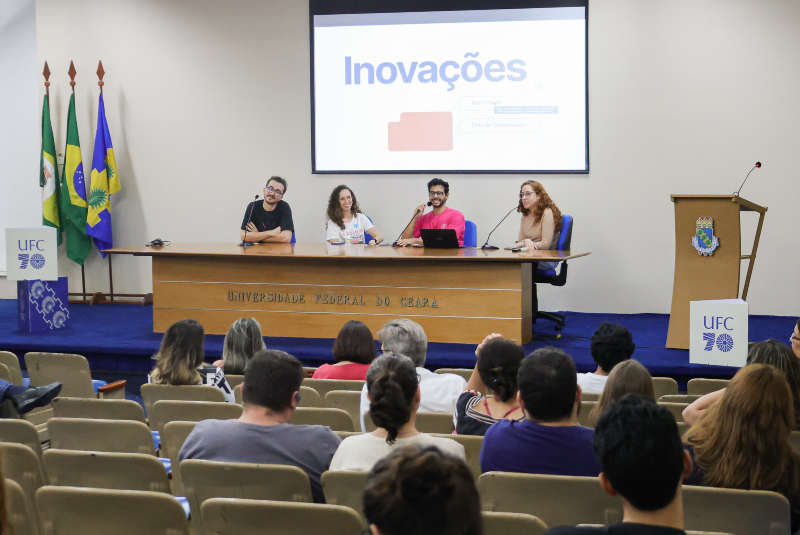 Imagem: A imagem mostra um momento do 1º Encontro da Rede de Comunicadores da UFC, realizado no auditório da Reitoria da Universidade Federal do Ceará (UFC). No palco, quatro comunicadores estão sentados à mesa principal, participando de uma mesa-redonda ou painel sobre inovações na comunicação. Atrás deles, um telão exibe a palavra “Inovações” em destaque, junto a uma imagem de uma pasta digital, indicando o tema da apresentação.  À esquerda da imagem, veem-se novamente as bandeiras do Brasil, do estado do Ceará e da UFC, reforçando o caráter institucional do evento. A mesa possui o nome da universidade gravado na parte frontal, e o púlpito ao lado também exibe o logotipo dos 70 anos da UFC.  O público, composto por profissionais e estudantes da área de comunicação, está sentado nas cadeiras do auditório, atento aos palestrantes. O ambiente é informal, porém profissional, com clima de troca de experiências e atualização sobre práticas comunicacionais.  O evento demonstra o esforço da universidade em fortalecer sua rede interna de comunicação, promovendo integração, inovação e compartilhamento de boas práticas entre os comunicadores da instituição.