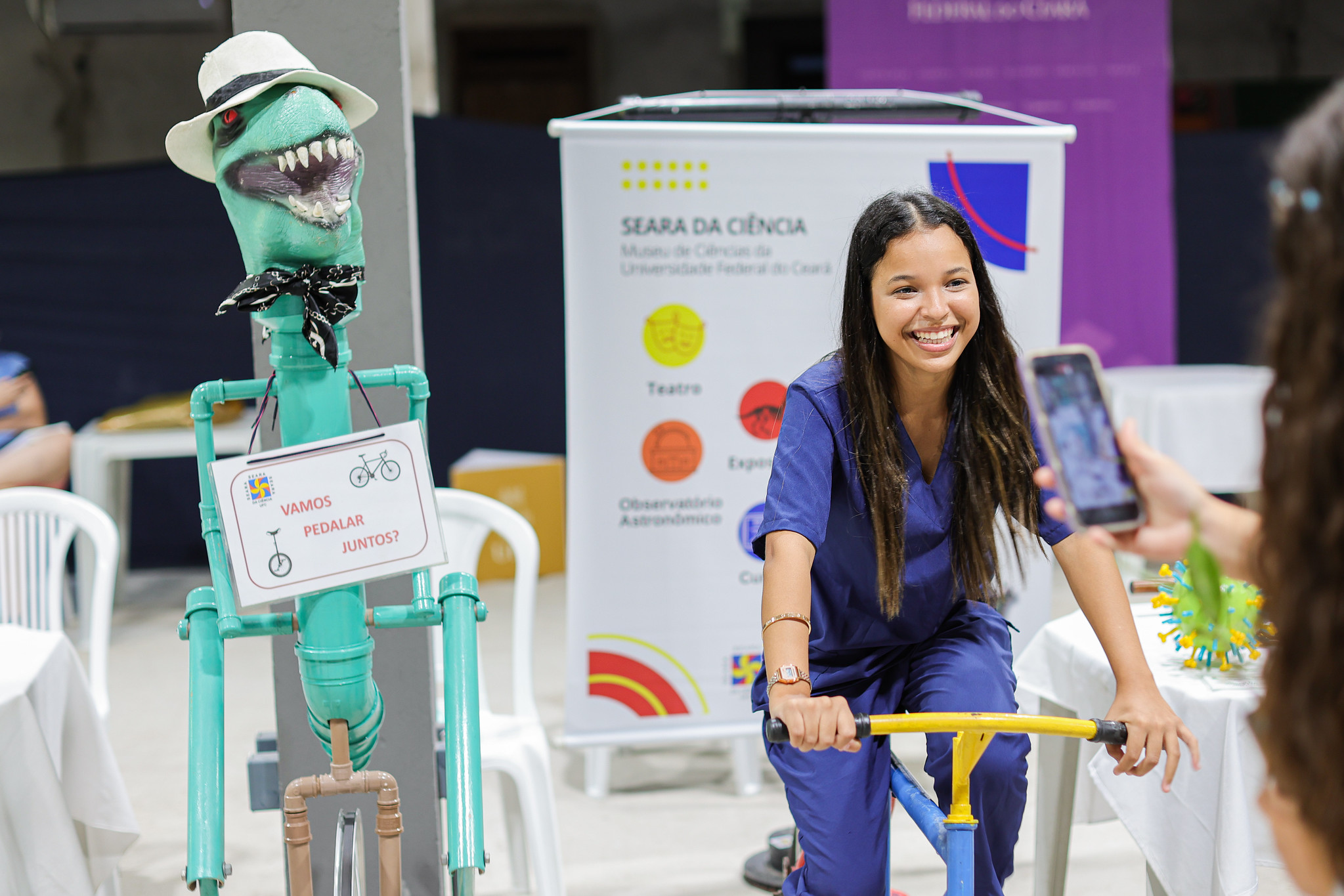 Imagem: Jovem sorridente, vestida com uniforme azul, pedala uma bicicleta estacionária em uma atividade interativa da Seara da Ciência, da Universidade Federal do Ceará (UFC). Ao lado dela, há uma figura lúdica feita com canos coloridos, com cabeça de dinossauro, chapéu branco e lenço no pescoço, segurando uma placa com os dizeres “Vamos pedalar juntos?”. Ao fundo, um banner com informações sobre o museu destaca atividades como teatro, espaço de experimentos e observatório astronômico. Outra pessoa registra o momento com um celular. O ambiente é claro e descontraído.