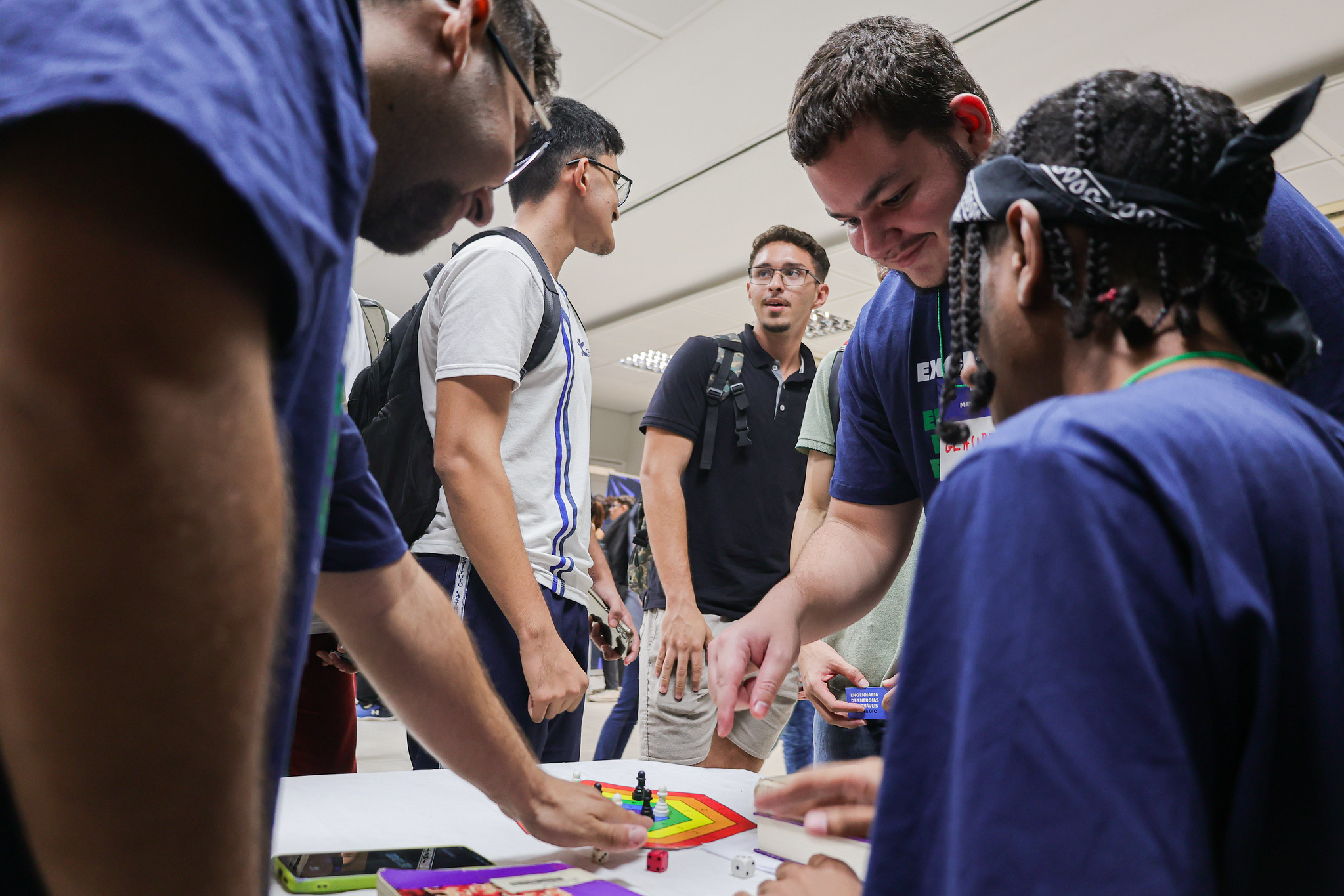 Imagem: Grupo de jovens interage em torno de uma mesa com um tabuleiro colorido e dados, em uma atividade lúdica organizada durante evento da Universidade Federal do Ceará (UFC). Dois participantes, com camisetas do evento, movimentam peças no jogo, enquanto outros observam e conversam. O ambiente é fechado e bem iluminado, com teto branco e luminárias. Ao fundo, mais estudantes circulam, muitos com mochilas nas costas, indicando que estão visitando os estandes. A cena transmite um clima descontraído de aprendizado e integração.