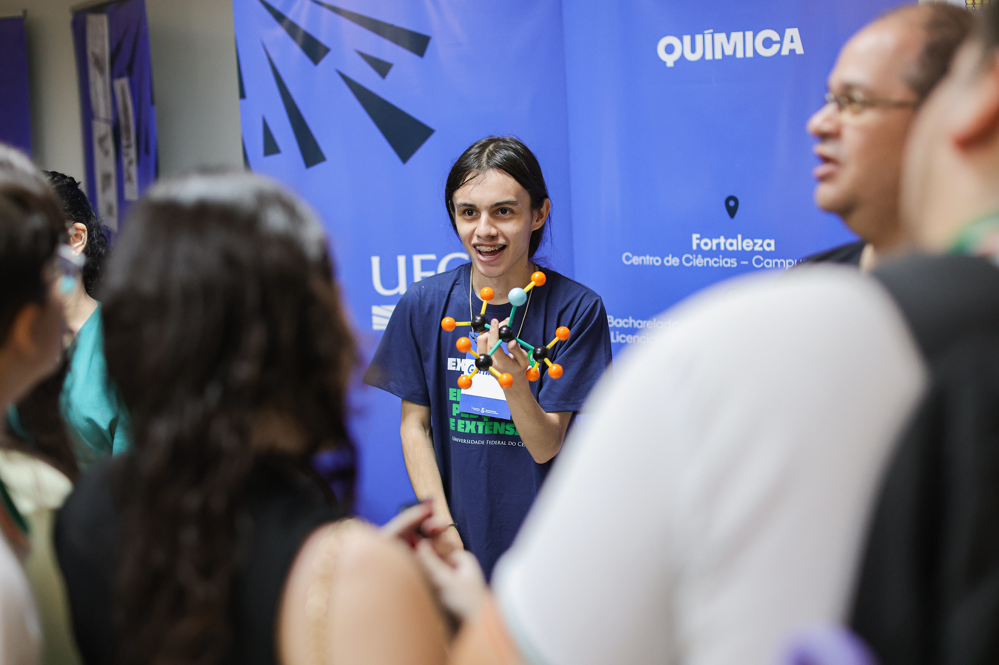 Imagem: Estudante do curso de Química da Universidade Federal do Ceará (UFC) sorri enquanto apresenta um modelo molecular feito com hastes e esferas coloridas. Ele está diante de um banner azul com a palavra "Química" em destaque e informações sobre o curso no Campus Fortaleza. À frente dele, há um grupo de pessoas ouvindo atentamente a explicação. A cena ocorre em um ambiente fechado, provavelmente durante um evento de divulgação científica ou feira acadêmica.