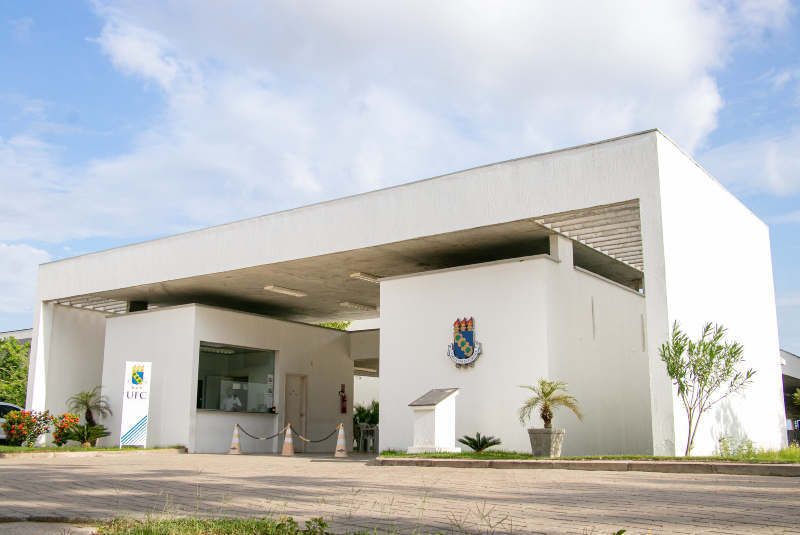Imagem: A imagem mostra a fachada principal do campus da Universidade Federal do Ceará (UFC) em Russas. A construção tem um design moderno e minimalista, com linhas retas e predominância da cor branca. A estrutura principal é um pórtico retangular que cobre a guarita de entrada, com grandes pilares que sustentam o teto.  Na parte frontal, há o brasão da UFC fixado na parede e uma placa informativa logo abaixo. Ao lado, um totem vertical exibe o logotipo da universidade. A entrada possui uma guarita com vidraça para controle de acesso. A área ao redor é bem cuidada, com vasos de plantas ornamentais, arbustos, pequenas palmeiras e flores, conferindo um aspecto acolhedor ao ambiente. O céu está parcialmente nublado, e a iluminação natural valoriza ainda mais a arquitetura limpa e funcional da entrada do campus
