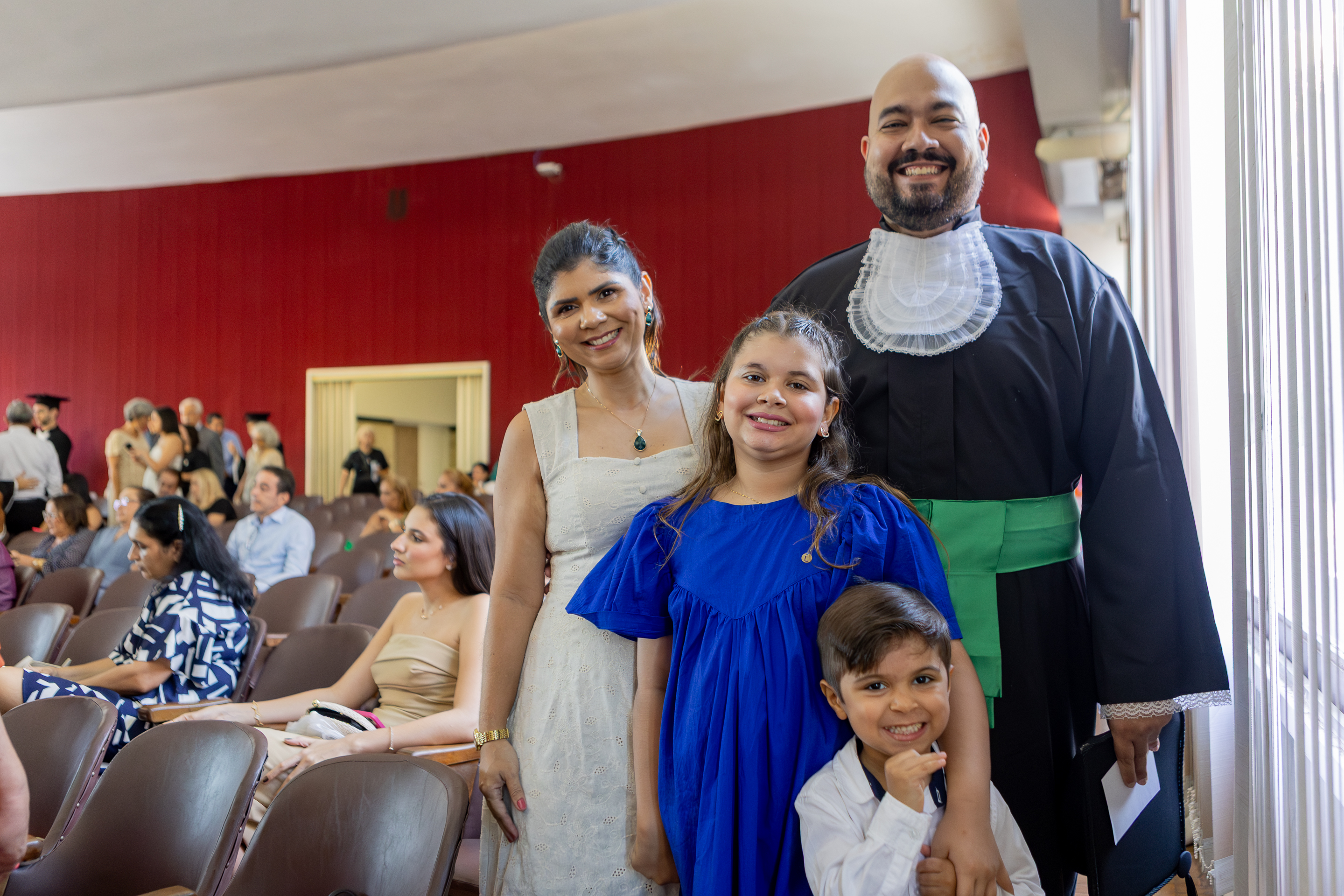Imagem: homem calvo e sorridente usa beca preta de formatura com faixa verde na cintura e adorno branco rendado no peito. Ele está em pé ao lado de sua família: uma mulher de vestido claro, uma menina usando vestido azul e um menino pequeno sorrindo e segurando o braço da irmã. Eles estão em um auditório, cercados por outras pessoas sentadas e em pé, em clima de comemoração. A luz natural entra pelas janelas à direita, iluminando o grupo. A cena transmite alegria e união familiar em um momento especial de conquista.