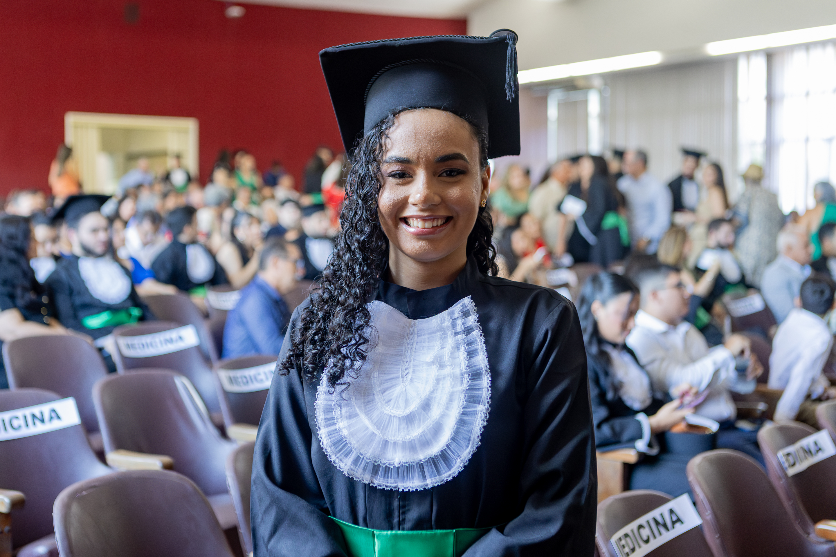 Imagem: mulher jovem parda sorri enquanto posa para a foto usando uma beca preta de formatura, com um adorno rendado branco no peito e uma faixa verde na cintura. Ela também usa um capelo preto (chapéu de formatura). O cenário é um auditório lotado, com várias pessoas ao fundo, algumas também usando becas e outras roupas formais. As cadeiras ao redor estão identificadas com a palavra “MEDICINA”, indicando que a cerimônia é de formatura de um curso de Medicina. A atmosfera é alegre e festiva.