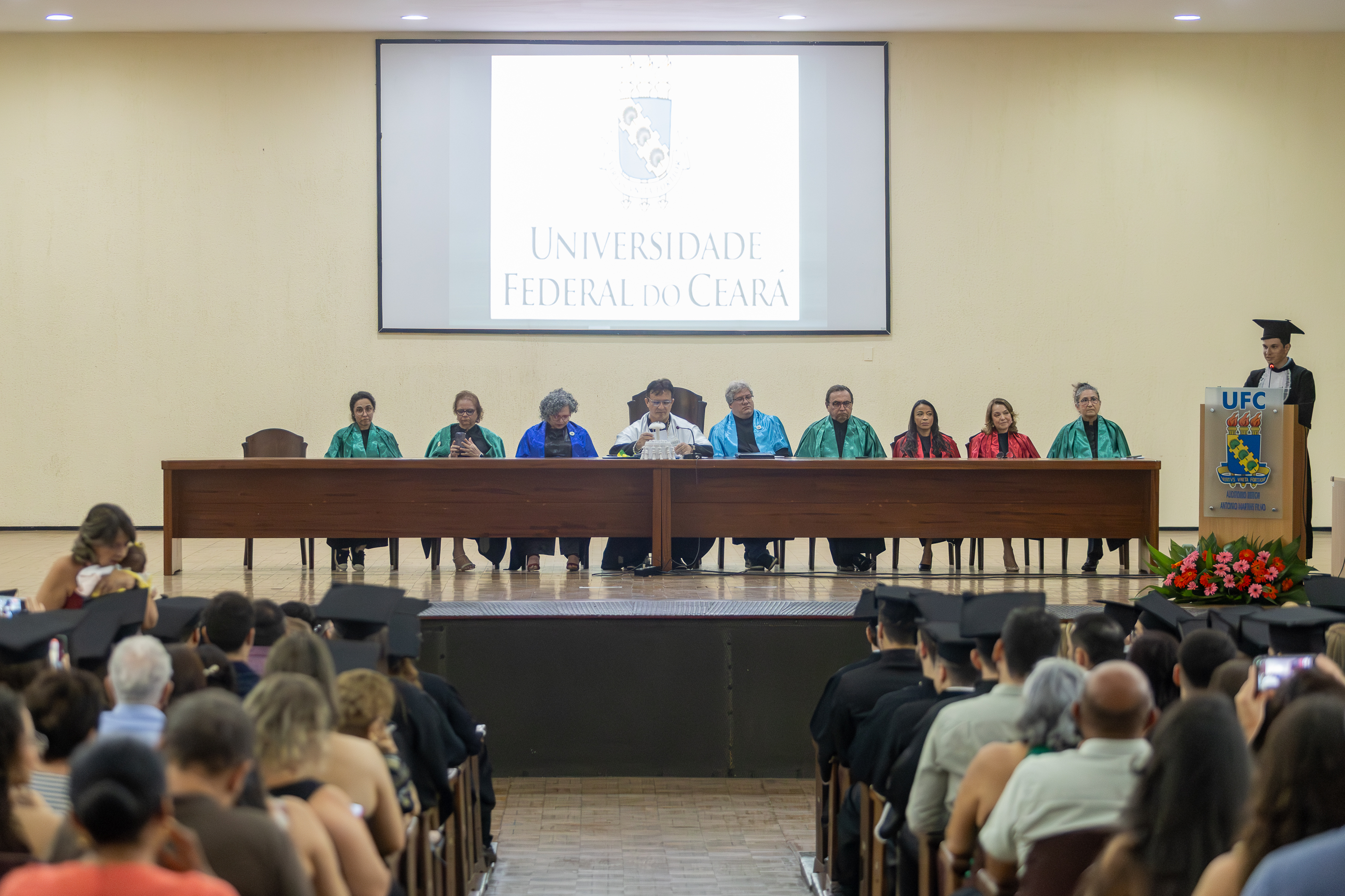 Imagem: auditório com diversas pessoas assistindo a uma cerimônia de formatura. Ao fundo, um palco com uma longa mesa onde estão sentados professores e autoridades acadêmicas da Universidade Federal do Ceará, identificada pelo logotipo projetado em uma grande tela ao fundo. As vestes dos membros da mesa são coloridas, indicando diferentes áreas do conhecimento. À direita, um formando em pé, de beca e capelo, faz um discurso no púlpito com o brasão da universidade e um arranjo de flores na base. A atmosfera é solene e festiva.