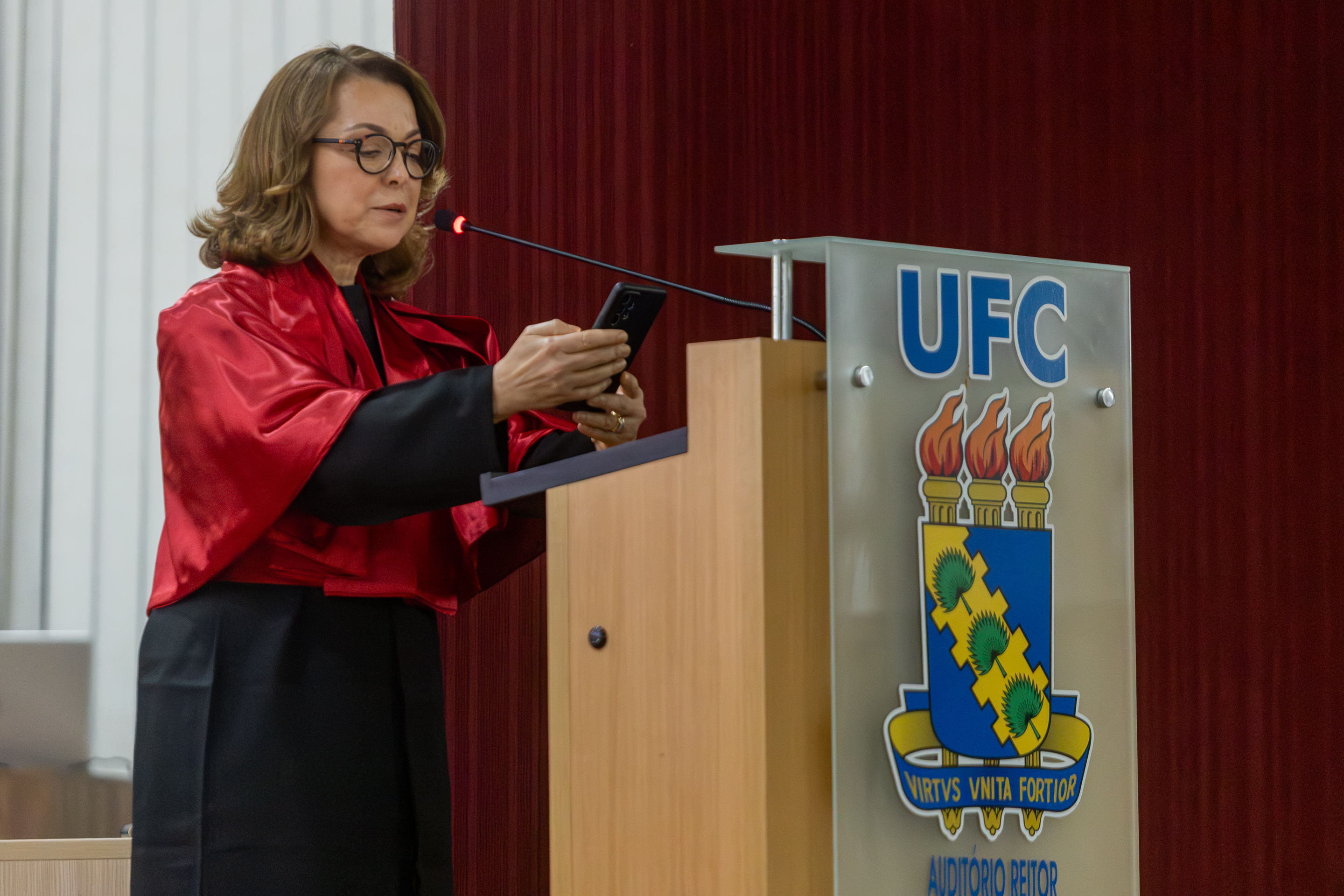 Imagem: mulher branca, de cabelos castanhos claros ondulados e de óculos, vestindo uma beca preta com faixa vermelha, fala ao microfone em um púlpito durante uma cerimônia. Ela segura um celular enquanto se dirige ao público. No púlpito está o brasão da Universidade Federal do Ceará (UFC) com três tochas acesas e a frase em latim “Virtvs Vnita Fortior”. Ao fundo, a parede é revestida com cortina clara e painel vermelho.