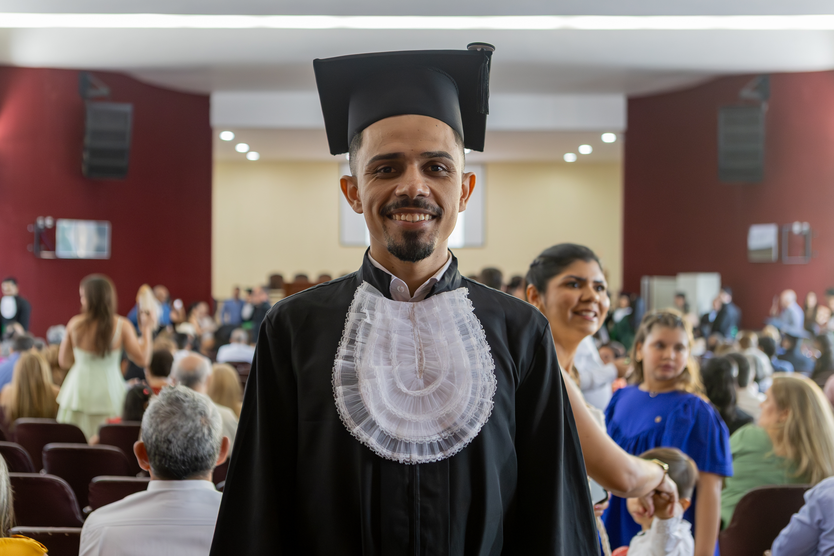 Imagem: homem jovem negro, de cavanhaque, está sorrindo, vestindo uma beca preta e um capelo (chapéu de formatura) preto com um adorno branco rendado no peito, típico de cerimônias de formatura no Brasil. Ele está em pé em um auditório cheio de pessoas, sugerindo que está participando de sua cerimônia de formatura. Ao fundo, há várias pessoas sentadas e em pé, com destaque para uma mulher sorridente e uma criança de vestido azul. O ambiente é iluminado e festivo.