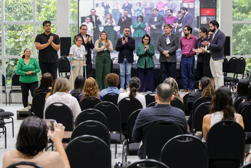 Imagem: A imagem mostra um momento do Fórum Nordeste de Doenças Tropicais Negligenciadas, realizado no Centro de Convivência da Universidade Federal do Ceará (UFC), no Campus do Pici. No palco, está um grupo de 13 pessoas, formado por organizadores, pesquisadores e autoridades da área da saúde e da ciência, todos posicionados de pé, lado a lado, e aplaudindo. Algumas pessoas no palco também demonstram sorrisos e expressões de satisfação. Ao fundo, há um telão grande exibindo uma imagem semelhante à da cena ao vivo, provavelmente uma transmissão ou foto do próprio evento. A parede de vidro atrás do palco permite ver a vegetação verde do exterior, criando um ambiente iluminado e natural. Na plateia, diversas pessoas estão sentadas em cadeiras pretas, voltadas para o palco, acompanhando atentamente. Uma participante à frente está registrando o momento com seu celular. A atmosfera transmite um evento acadêmico e institucional, voltado à discussão e visibilidade de temas científicos importantes e muitas vezes esquecidos pela saúde pública, como as doenças tropicais negligenciadas