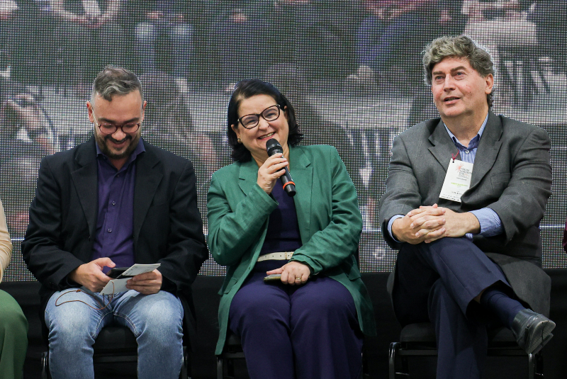 Imagem: A imagem mostra um momento descontraído durante uma mesa de discussão ou painel no Fórum Nordeste de Doenças Tropicais Negligenciadas, realizado no Centro de Convivência da Universidade Federal do Ceará (UFC), no Campus do Pici.  Três pessoas estão sentadas no palco, em destaque, provavelmente pesquisadores, professores ou gestores da área da saúde e ciência. O clima é leve e acolhedor: a mulher ao centro, que está com um microfone na mão, sorri enquanto fala, transmitindo simpatia e envolvimento com o público. Ela usa um conjunto verde e azul escuro e está acompanhada por dois homens — o da esquerda segura papéis e sorri olhando para baixo, enquanto o da direita observa atentamente, com postura relaxada e crachá pendurado no pescoço.  Ao fundo, um telão de LED exibe uma imagem do evento, ampliando a experiência para a plateia. O ambiente comunica um encontro formal, porém acessível, promovendo o diálogo técnico e científico de forma engajada