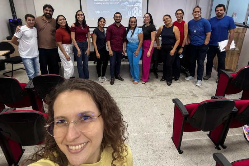 Imagem: A imagem retrata a comissão setorial do Processo Estatuinte da Universidade Federal do Ceará (UFC) no Campus de Crateús. O grupo é formado por cerca de 16 pessoas, entre mulheres e homens, que estão organizados em pé, lado a lado, em frente a um telão de projeção. No telão, é possível visualizar parte de uma apresentação, que reforça o caráter institucional do encontro.  Em primeiro plano, há uma mulher sorrindo que tira uma selfie com o grupo ao fundo, trazendo um tom de descontração e proximidade à imagem. O ambiente é uma sala de aula ou auditório, com cadeiras vermelhas e pretas organizadas, piso em granito claro e iluminação bem distribuída.  O grupo expressa diversidade, união e compromisso, representando diferentes segmentos da comunidade acadêmica — docentes, técnicos e discentes — reunidos no contexto democrático e participativo do Processo Estatuinte da UFC no interior do estado