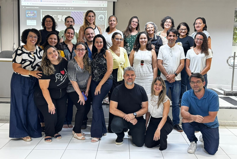 Imagem: A imagem mostra um grupo de aproximadamente 26 pessoas, composto por homens e mulheres, reunidos em um ambiente interno, possivelmente uma sala de reuniões ou auditório. Todos estão posando para a foto, muitos sorrindo, transmitindo um clima de colaboração e acolhimento.  Ao fundo, há um telão com projeções que fazem referência ao Processo Estatuinte da Universidade Federal do Ceará (UFC), evidenciado por elementos gráficos e textos parciais visíveis na apresentação. As pessoas estão organizadas em três fileiras: uma em pé, uma agachada ou ajoelhada à frente e outra no meio, ligeiramente abaixada.  O grupo representa a comissão setorial do Processo Estatuinte da UFC, formada por docentes, técnico-administrativos e discentes, demonstrando diversidade de gênero, estilos e faixas etárias. O ambiente tem paredes claras, piso branco e uma atmosfera que reforça o caráter institucional, democrático e participativo do momento registrado