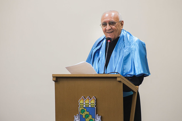 Imagem: um homem idoso, de pele clara, cabelos curtos e brancos, usa óculos de grau e veste beca preta com faixa azul. Ele está em pé atrás de um púlpito de madeira com o brasão da Universidade Federal do Ceará. Sorri enquanto segura folhas de papel com a mão esquerda e fala ao microfone com espuma protetora preta e vermelha. O fundo da imagem é claro e liso.