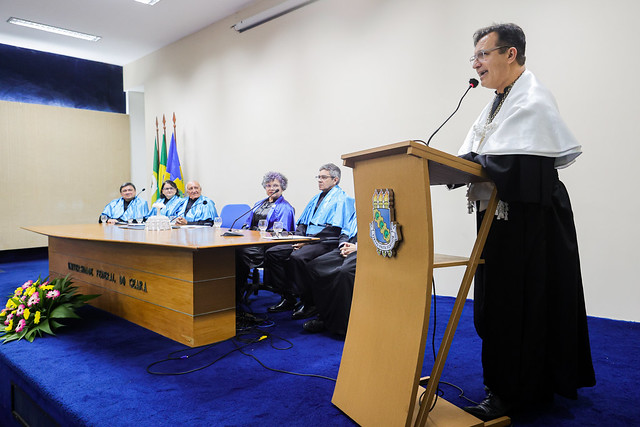 Imagem: um homem de pele clara, com óculos de grau e beca preta com capa branca, fala em um púlpito de madeira com o brasão da Universidade Federal do Ceará. Ele segura o microfone com espuma preta e vermelha. À esquerda, uma mesa com seis pessoas sentadas, quatro homens e duas mulheres, todos em beca preta com faixa azul, observam a fala. A mesa de madeira tem a inscrição "Universidade Federal do Ceará". No canto inferior esquerdo, um arranjo de flores coloridas. Ao fundo, bandeiras do Brasil, do Ceará e da UFC. O ambiente é uma sala com carpete azul e paredes claras.