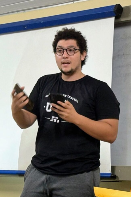Imagem: Foto de um jovem de pele clara, cabelos cacheados e curtos, barba rala e usando óculos de grau redondos. Ele veste uma camiseta preta com o logotipo da Universidade Federal do Ceará (UFC) e calça esportiva cinza. Está em pé, em uma sala de aula, diante de um quadro branco, segurando um celular em cada mão e aparenta estar falando para um público. Ao fundo, parte de um projetor e anotações em um quadro