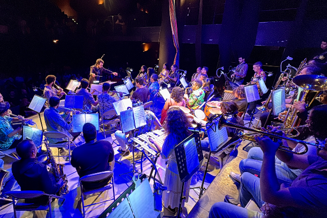 Imagem: Foto tirada do palco durante uma apresentação musical. Em destaque, uma grande formação de músicos está disposta em semicírculo, todos sentados e tocando instrumentos como saxofones, trompetes, trombones, flautas, percussão e teclado. No centro, uma mulher com gestos expressivos rege a banda sinfônica. O palco está iluminado por luzes azuladas, enquanto o público, ao fundo, aparece em penumbra, assistindo ao espetáculo em um teatro. As partituras nos suportes e a variedade de instrumentos mostram a complexidade e o preparo da apresentação.