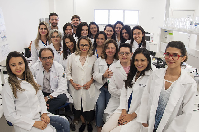 Imagem: foto em ambiente de laboratório, bem iluminado, com paredes brancas, bancada com vidrarias e microscópios ao fundo. No centro da imagem, um grupo de 20 pessoas posa sorridente para a câmera. A maioria veste jaleco branco, indicando atuação na área da saúde ou científica. Na primeira fileira, sete pessoas estão sentadas ou agachadas; ao centro, está uma mulher de jaleco, com óculos e cabelos soltos. As demais pessoas, homens e mulheres, estão em pé atrás dela, dispostas em fileiras e bem próximas umas das outras, demonstrando união e espírito de equipe.