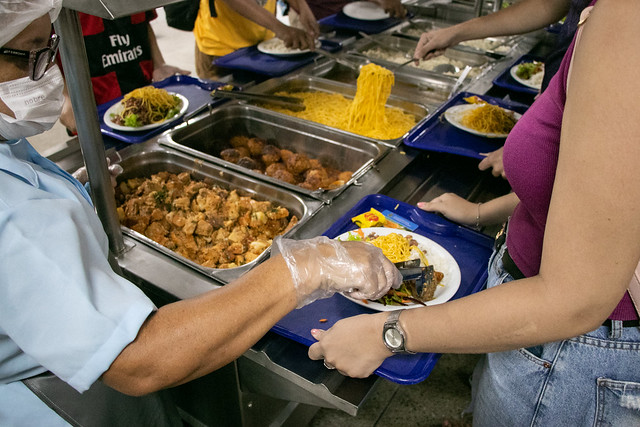 Imagem: Pessoas em fila se servem em um restaurante universitário. Bandejas de inox cheias de alimentos, como macarrão, legumes e pratos preparados, estão dispostas sobre o balcão. Uma funcionária, usando touca, máscara e luvas descartáveis, serve a refeição no prato de uma estudante que segura uma bandeja azul. Outras pessoas aguardam para se servir, cada uma com sua bandeja.