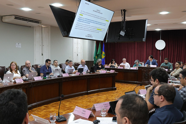 Imagem: reunião em uma sala ampla, com várias pessoas sentadas em torno de uma grande mesa em formato de “U”. Todos estão voltados para o centro e muitos utilizam notebooks e papéis. No teto, dois telões exibem uma apresentação com texto. Ao fundo, há cortinas vermelhas fechadas, duas bandeiras — do Brasil e do Ceará — e uma parede clara. O ambiente é de deliberação e discussão formal.